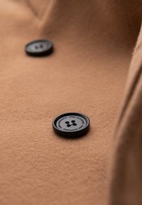 Close-up af to sorte knapper med tekst på et brunt uldfrakke-stof, der fremhæver frakkens tekstur og knapdesign.