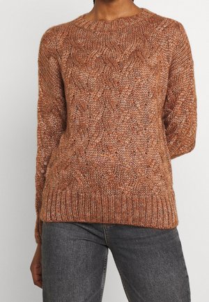 Person iført en rustfarvet kabelstrikket sweater med lange ærmer og ribbede manchetter, kombineret med mørkegrå jeans.