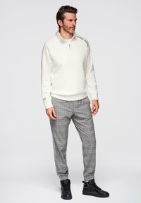 Sudadera blanca con cremallera, pantalones a cuadros grises con rayas negras y zapatillas negras, usados por un hombre con cabello corto y barba.