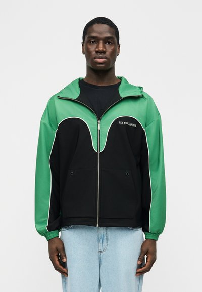 Άνδρας που φοράει μαύρο και πράσινο τζάκετ με φερμουάρ και το κείμενο "LES BENJAMINS", συνδυασμένο με ανοιχτόχρωμα μπλε τζιν, στέκεται μπροστά σε απλό φόντο.