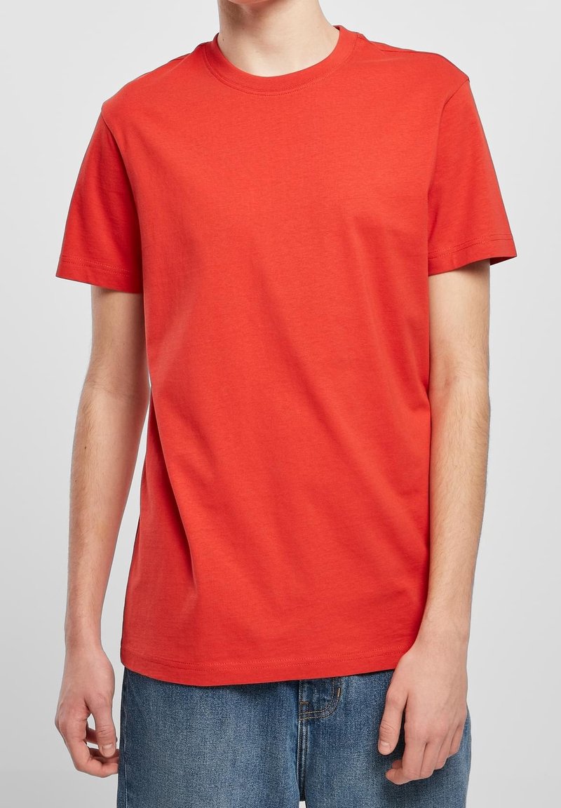 Personne portant un t-shirt rouge uni à manches courtes et un jean bleu, debout devant un fond gris clair.