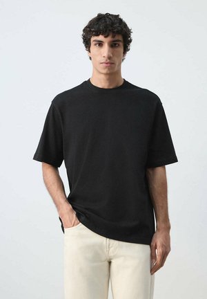 Junger Mann mit lockigem dunklem Haar, der ein schlichtes schwarzes T-Shirt und hellbeige Hose trägt, steht mit einer Hand in der Tasche vor einem einfarbigen Hintergrund.