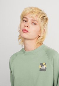 Personne avec les cheveux blonds courts portant un sweat à capuche vert clair avec un logo floral North Face sur la poitrine, sur fond uni.