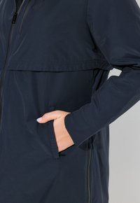 Námornícky voděodolný kabát s kapsou na zip, měkkou tkaninou a uvolněným střihem. Obsahuje klopu na přední straně a dlouhé rukávy.