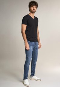 Camisa negra de cuello en V, de tejido de peso medio, combinada con jeans entallados de color azul claro y zapatillas blancas con acentos grises, sobre un fondo neutro.