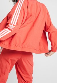 Červená lehká bunda s bílým designem tří pruhů na rukávech, elastickým pasem a logem Adidas na levé paži.