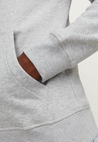 Sudadera gris con un tejido texturizado, que presenta un bolsillo canguro y puños de canalé. La manga muestra un tono de piel en contraste.