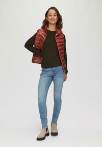 Rote Steppweste über einem dunkel strukturierten Sweater, kombiniert mit hellblauen Jeans und beigen Knöchelstiefeln. Einfaches Design ohne sichtbare Muster.