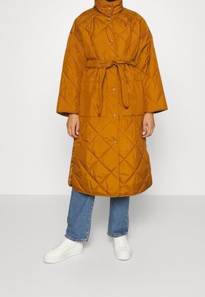 Długi pikowany płaszcz w kolorze musztardowym z przednimi zatrzaskami i paskiem w talii, noszony na niebieskich dżinsach i białych butach sportowych.