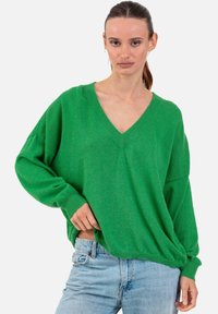Femme portant un pull ample à col en V vert à manches longues et un jean en denim bleu clair, debout devant un fond uni.