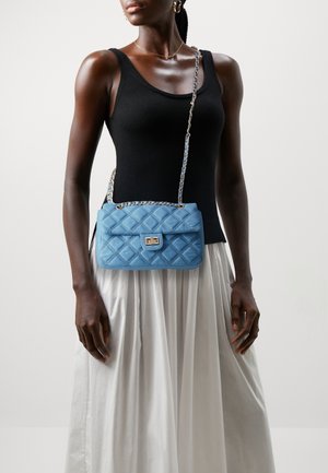 Sac à main matelassé bleu clair avec un fermoir et une bandoulière chaîne en métal argenté, présentant une texture douce et une forme rectangulaire, porté avec un haut noir.