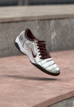 Pantof sport Nike argintiu și burgundy cu șireturi, plutind deasupra unei suprafețe din piatră roz și cu un perete de piatră gri în fundal.