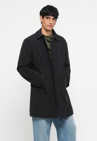 Μαύρο, ελαφρύ παλτό με ίσια γραμμή, μακριά μανίκια, πλαϊνές τσέπες και κολάρο, φορεμένο πάνω από πουκάμισο με πράσινο σχέδιο.