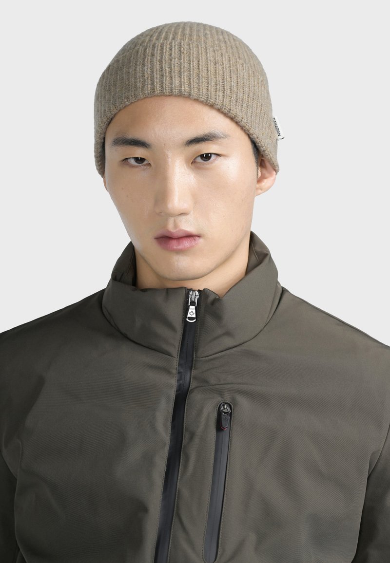 Gorro de canalé marrón, usado con una chaqueta azul oscuro con cremallera que presenta un cuello alto y un bolsillo en el pecho con cremallera. Textura de tela suave.