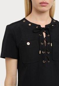 Černá košile s krátkými rukávy, jež má zlaté očka a šněrovací design, dvě přední kapsy a hladkou texturu látky.