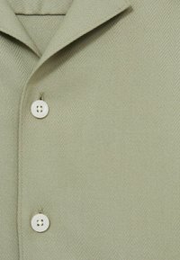 Camisa verde claro con textura de espiga, cuello puntiagudo y tres botones blancos en la tapeta.