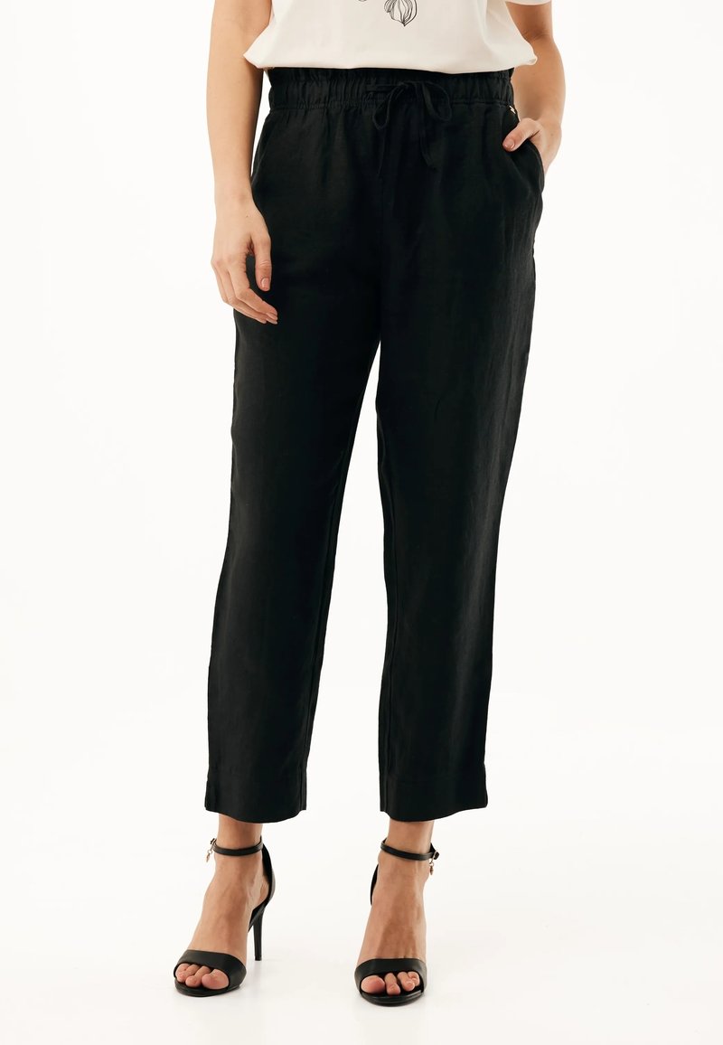 Vrouw draagt zwarte high-waist taps toelopende broek met elastische tailleband en zwarte hoge hak sandalen met enkelriempje, staat met één hand in de zak.