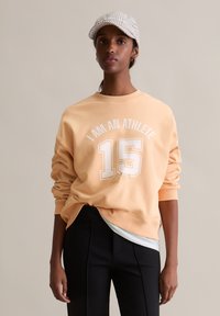 Brzoskwiniowy sweatshirt z napisem "JESTEM ATLETĄ" oraz "15" nadrukowanym na biało, luźny krój, ściągacze na rękawach, w zestawieniu z czarnymi spodniami pod lekką warstwą.