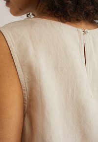 Top en lin beige avec un col rond et une subtile fente au dos, fermé par un bouton décoratif. Texture lisse avec des coutures minimales.