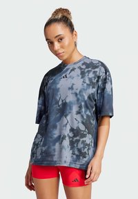 Naine, kellel on lokkis kukkel, kannab üliksuurt sinise ja musta sidrunimustri särki ning erksaid punaseid lühikesi pükse musta logoga, seismas ühtlase tausta ees.