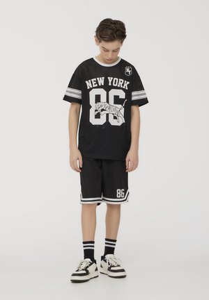 Maglia nera in rete con stampa "NEW YORK 86", grafica di tigre bianca, maniche a righe. Abbinata a pantaloni corti neri, dettagli bianchi e sneakers.