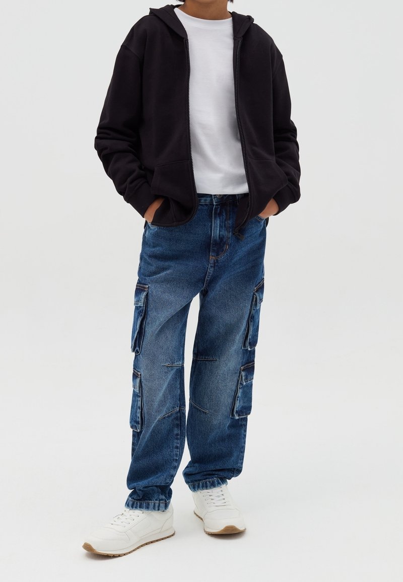 Persona che indossa una felpa con cappuccio nera con cerniera, maglietta bianca, jeans cargo blu con multiple tasche e scarpe da ginnastica bianche, in piedi con le mani nelle tasche.