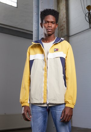 Giacca a vento gialla e blu navy con striscia bianca, chiusura con zip e cappuccio regolabile, abbinata a jeans blu chiaro.