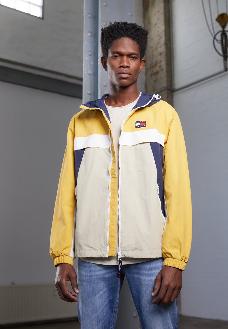 Giacca a vento gialla e blu navy con striscia bianca, chiusura con zip e cappuccio regolabile, abbinata a jeans blu chiaro.