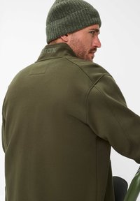 Felpa verde oliva, materiale in cotone, colletto alto, branding tonale sul retro, texture a costine. Indossata con un berretto lavorato a maglia abbinato.