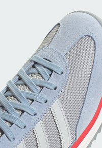 Light blue sneaker featuring suede and mesh materials, gray and white stripes, and red accents on the sole. Textured surface detail visible.