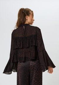 Mustat läpinäkyvät pusero, jossa on kerroksittain suunniteltu rakenne, sisältäen röyhelökoristeluja ja pitkät kellohihat. Kankaassa on hienovarainen leopardi-printti.