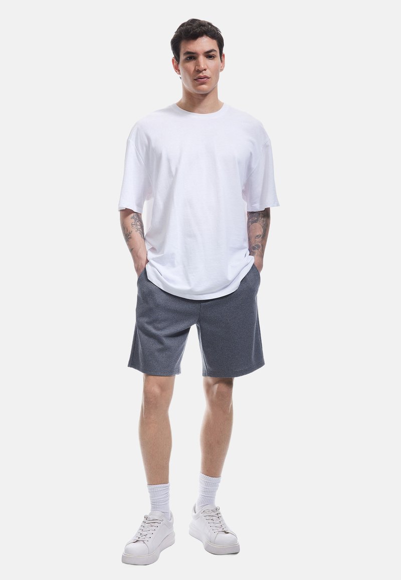 Weiße kurzärmlige T-Shirt, graue gemusterte Shorts und weiße Sneaker. Lässiger Schnitt, Baumwollmaterial, Hände in den Taschen, einfaches Design.