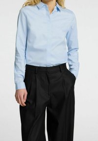Camicia azzurra chiaro con colletto, abbinata a pantaloni neri plissettati. La camicia ha maniche lunghe e una texture liscia.