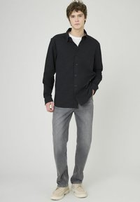 Jeune homme debout, portant une chemise noire à boutons, un t-shirt blanc en dessous, un jean gris et des chaussures beige clair, sur fond uni clair.