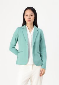 Ljusgrön blazer i mjukt tyg med skräddarsydd passform, kavajslag och två framfickor, bärs över en vit skjorta.
