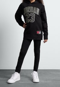 Černá mikina s kapucí s nápisem "JORDAN 23" tučným písmem, s štítkem na lemu, v kombinaci s černými legíny a bílými teniskami.