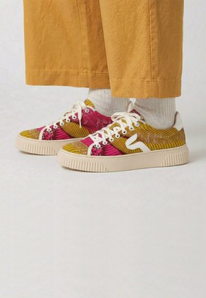 Baskets basses en tissu jaune et rose à motif, lacets blancs et semelles beige, portées avec des chaussettes blanches et un pantalon jaune moutarde.