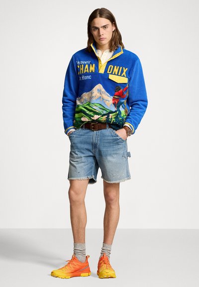Sudadera de forro polar azul con gráfico de montaña y texto, cuello amarillo y bolsillo. Combinada con pantalones cortos de denim desgastado y zapatillas deportivas naranja-amarillo.