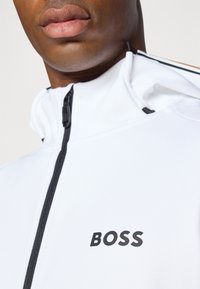 Weiße Zip-Jacke mit schwarzen Akzenten, ausgestattet mit einem hohen Kragen und einem schwarzen 'BOSS'-Logo auf der Vorderseite. Glattes, strukturiertes Material.