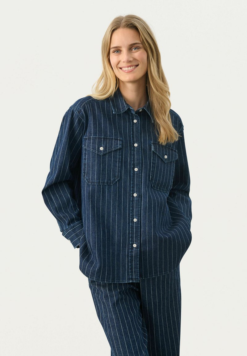 Camicia in denim con strisce bianche verticali, maniche lunghe, due tasche sul petto e chiusura frontale con bottoni. Base blu scuro, vestibilità rilassata.