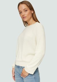 Helebeežik cardigan, mis on valmistatud tekstureeritud kudumiskangast, ümmarguse kaelusega ja pikkade varrukatega, koos helesiniste teksadega.