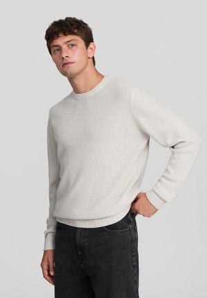 Maglione grigio chiaro a maglia con un motivo testurizzato, scollatura a girocollo e polsini a coste, abbinato a jeans scuri.