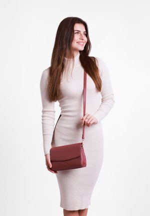 Burgundská kožená kabelka přes rameno s klopou, zlatými detaily a embosovaným logem. Modelka má na sobě upnuté krémové šaty s ribovaným vzorem.