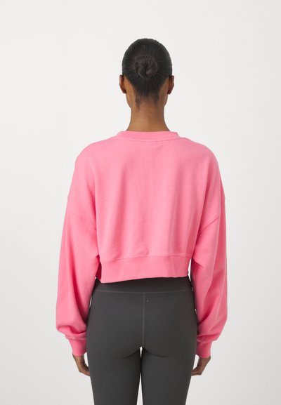 Rövidített rózsaszín pulóver, puha anyagból, esett vállakkal és bordás mandzsettákkal, fekete, testhez simuló leggingsszel párosítva.
