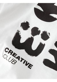 Valkoinen puuvillapaita, jossa on rohkeaa mustaa siveltimenveto-tekstiä, mukaan lukien "CREATIVE CLUB", sekä tekstuurinen graafinen muotoilu ja pehmeä viimeistely.