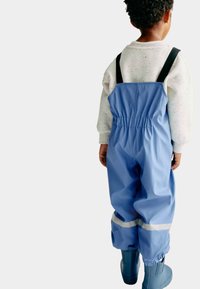 Salopette impermeabile blu chiaro con spalline nere regolabili, vita elasticizzata e dettagli riflettenti, abbinate a stivali di gomma blu.