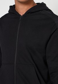 Czarna bluza z kapturem zapinana na zamek, wykonana z fakturowanej tkaniny, z kapturem, posiada kieszenie z przodu oraz tonacyjne akcenty na ramionach. Widoczny detal zamka błyskawicznego.