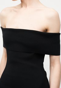 Γυναίκα με στενό μαύρο φόρεμα χωρίς τιράντες που αφήνει ακάλυπτους τους ώμους, το πάνω μέρος του στήθους και τα πάνω χέρια της, σε απλό φόντο.