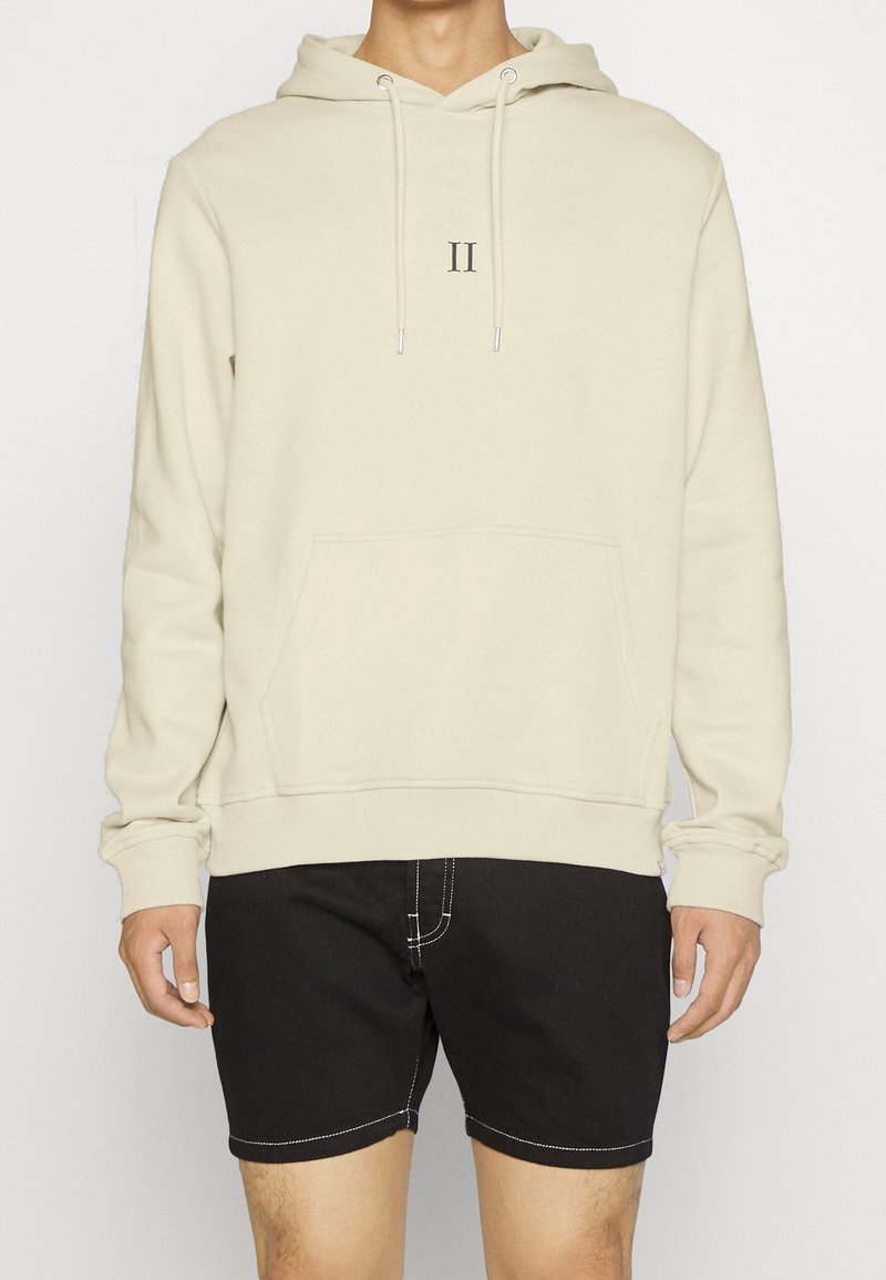 Personne portant un sweat à capuche beige clair avec une poche frontale et le logo "II", associé à un short noir avec des coutures blanches.