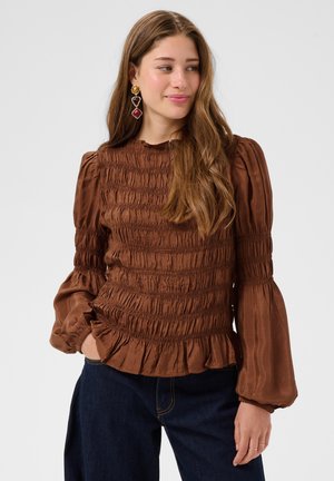 Femme aux longs cheveux bruns portant un haut à manches longues marron texturé et un jean bleu foncé, regardant sur le côté, les mains dans les poches.